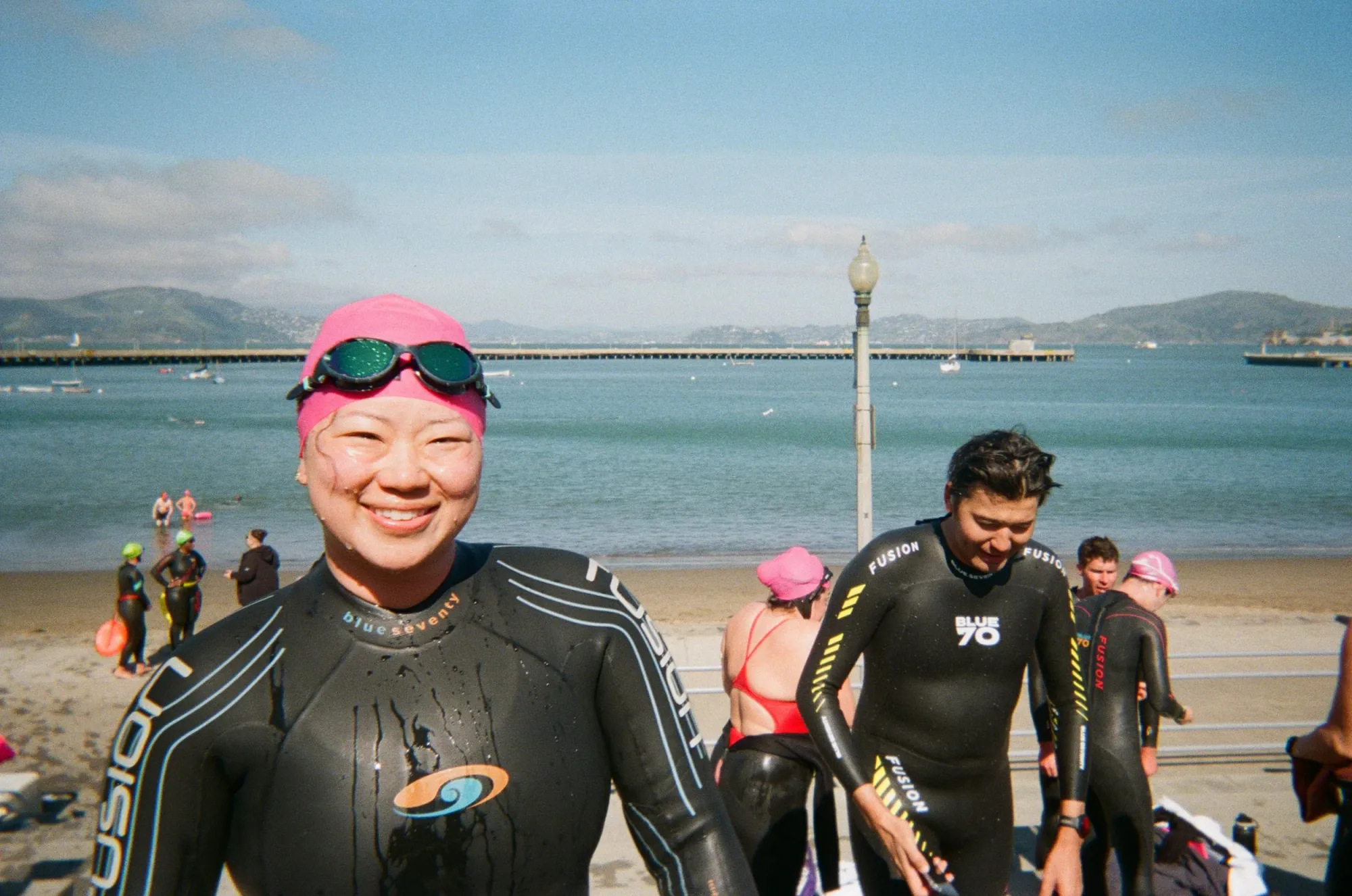 A club swimmer in a wetsuit and pink swim cap smiling on the beach after an open water swim, with other SF Tri Club swimmers and the bay behind her.